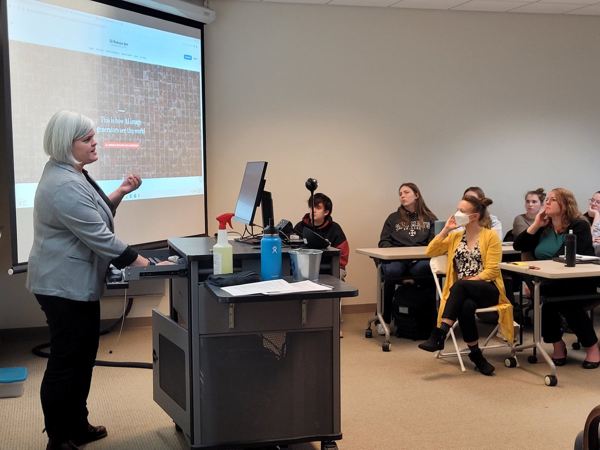 An instructor at a standing desk, speaking to a group of people seated in a classroom, with a projection screen and presentation behind her.